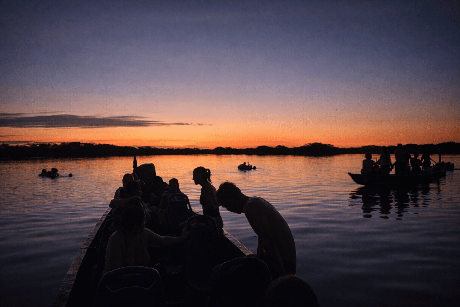 Siluetas de viajeros en canoas navegando el río Amazonas al atardecer, con aguas tranquilas y un horizonte iluminado.