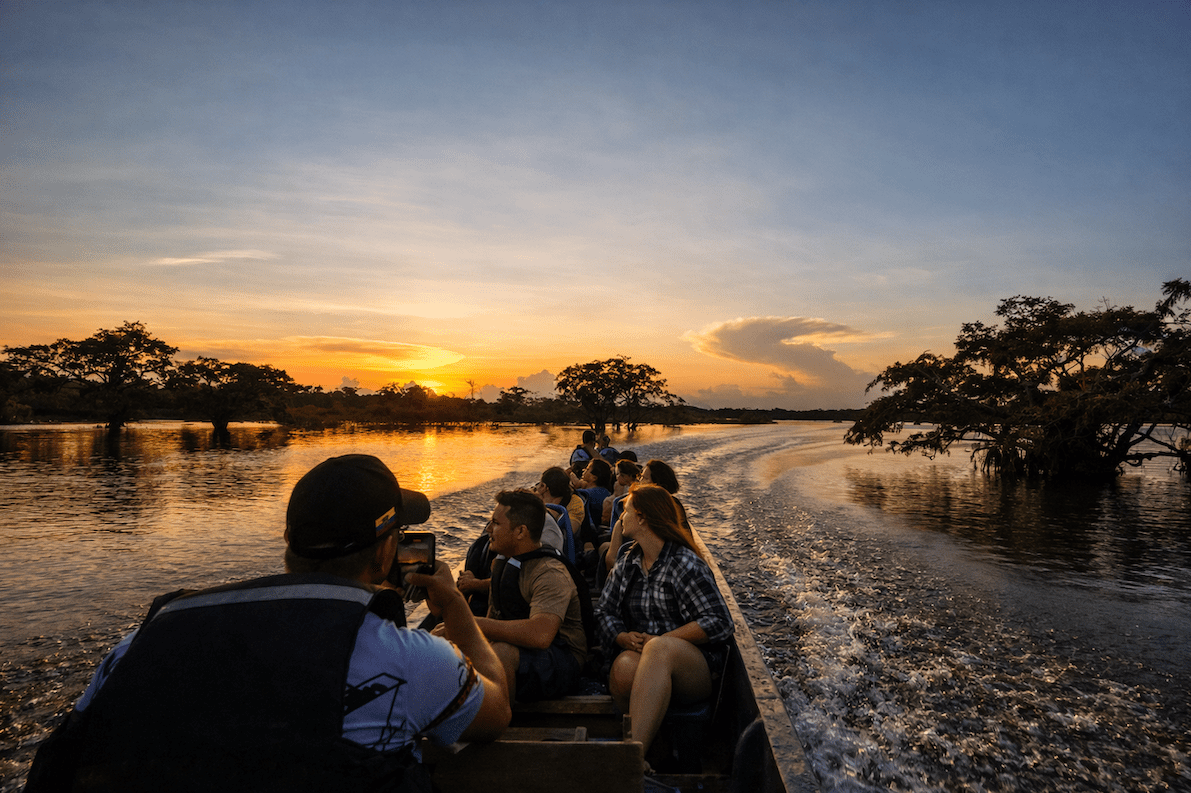 Huéspedes viajando en una canoa motorizada por un río amazónico al atardecer, rodeados de selva inundada.